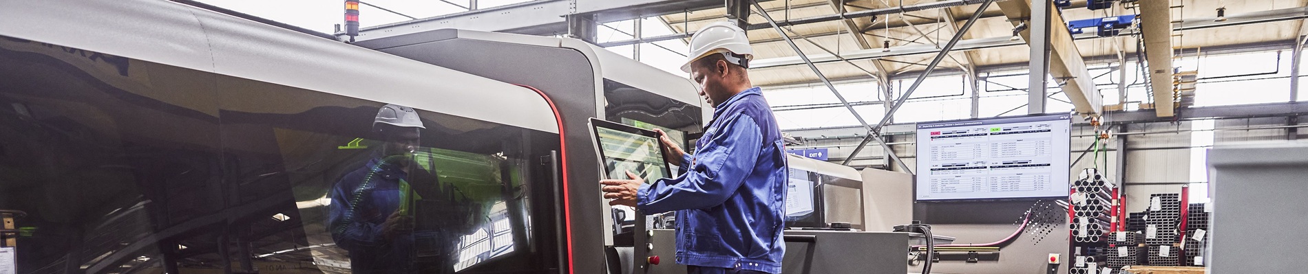 Ein Arbeiter in blauer Uniform und Helm bedient eine große Industriemaschine in einer Fabrik mit hohen Decken und einem digitalen Bildschirm.