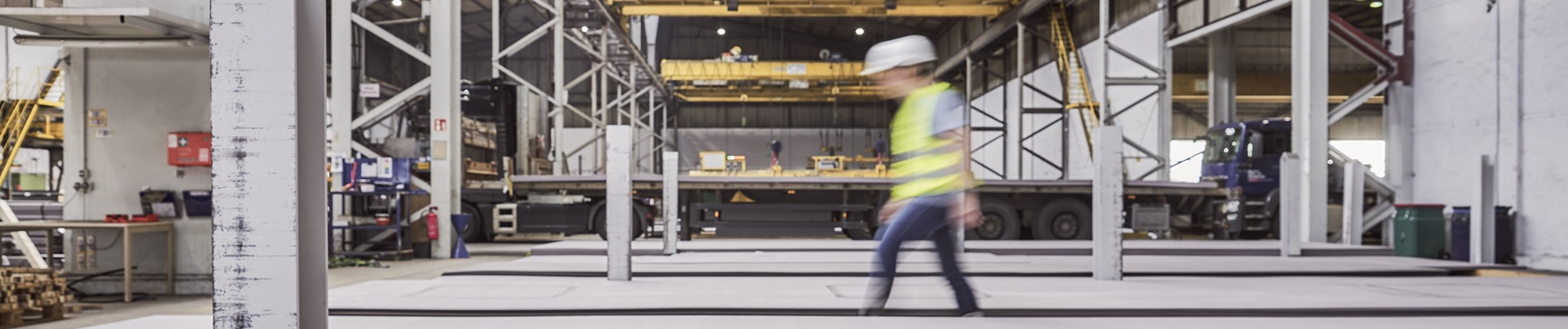 Eine Person mit Schutzhelm und Sicherheitsweste geht durch ein großes Industrie-Lagerhaus mit Metallträgern und Geräten.