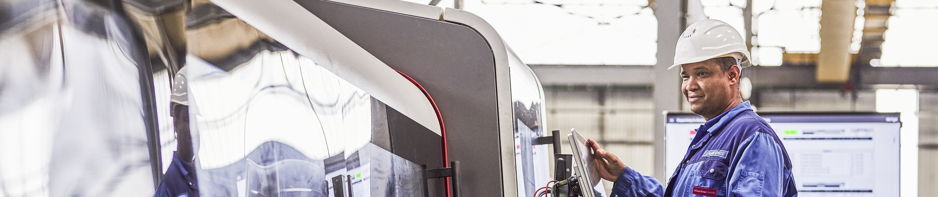 Arbeiter in blauer Arbeitskleidung und weißem Helm bedient Maschinen in einer industriellen Umgebung, im Hintergrund sind Bildschirme und Geräte zu sehen.Worker in blue overalls and white helmet operates machinery in an industrial setting, with screens and equipment visible in the background.