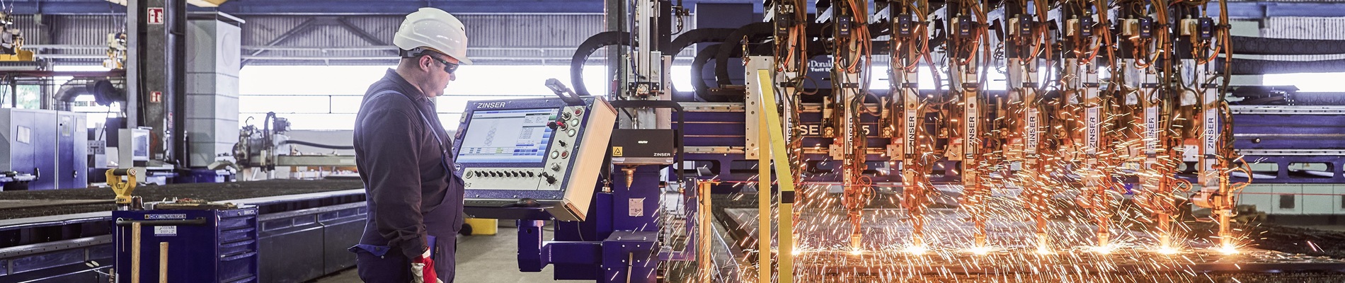 A worker in protective gear operates machinery in a factory, observing a process with bright sparks flying from a metal-cutting machine.