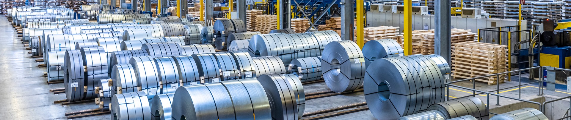 Großes Lagerhaus mit zahlreichen ordentlich gestapelten Coils und Holzpaletten unter heller Beleuchtung.