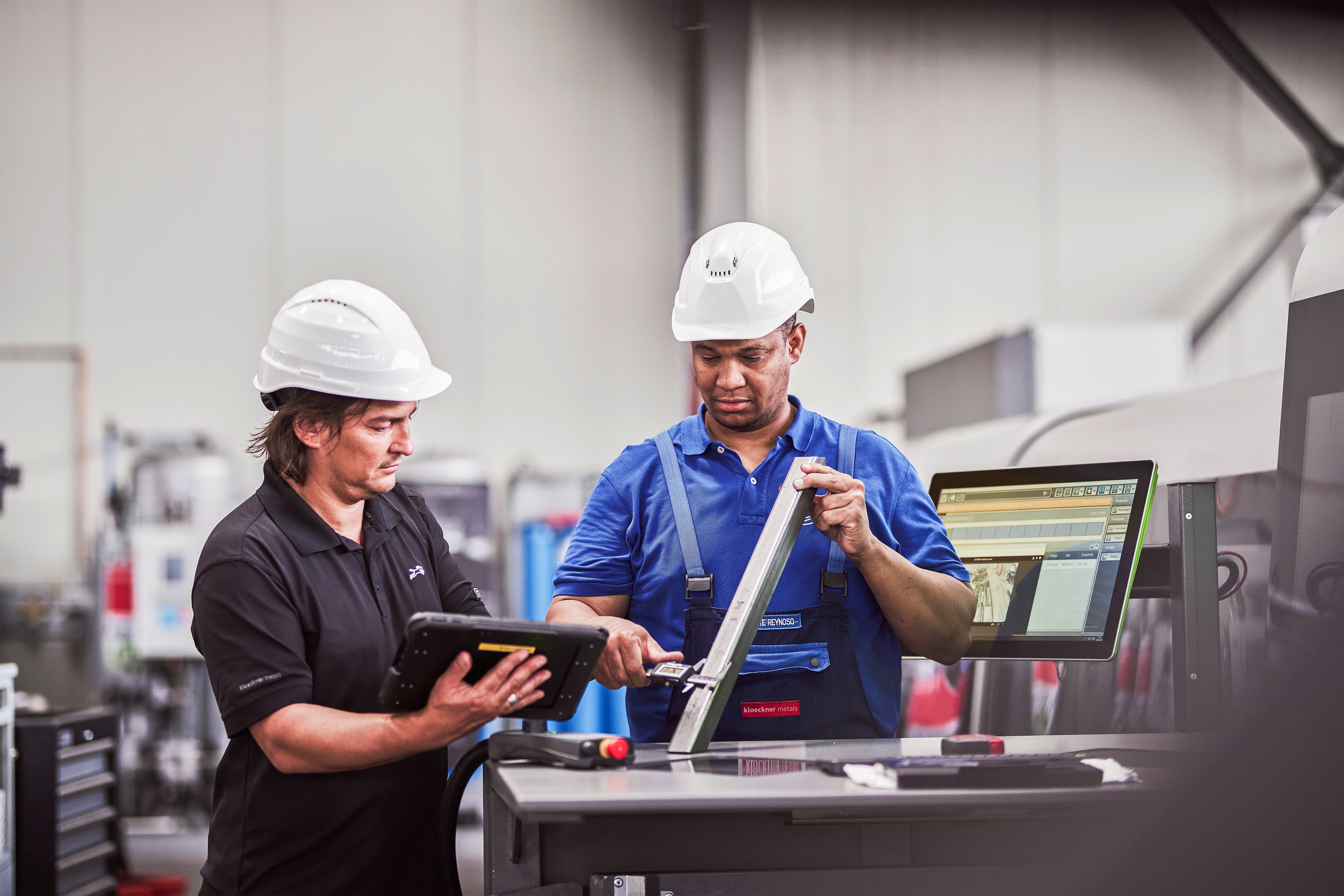Zwei Arbeiter in einer Produktionshalle tragen Schutzhelme und Arbeitskleidung. Einer hält ein Tablet in den Händen, während der andere mit einem Messgerät ein Metallprofil prüft. Im Hintergrund steht ein Computerbildschirm mit technischen Daten. Die Szene zeigt eine Zusammenarbeit zweier Mitarbeiter bei Kloeckner.