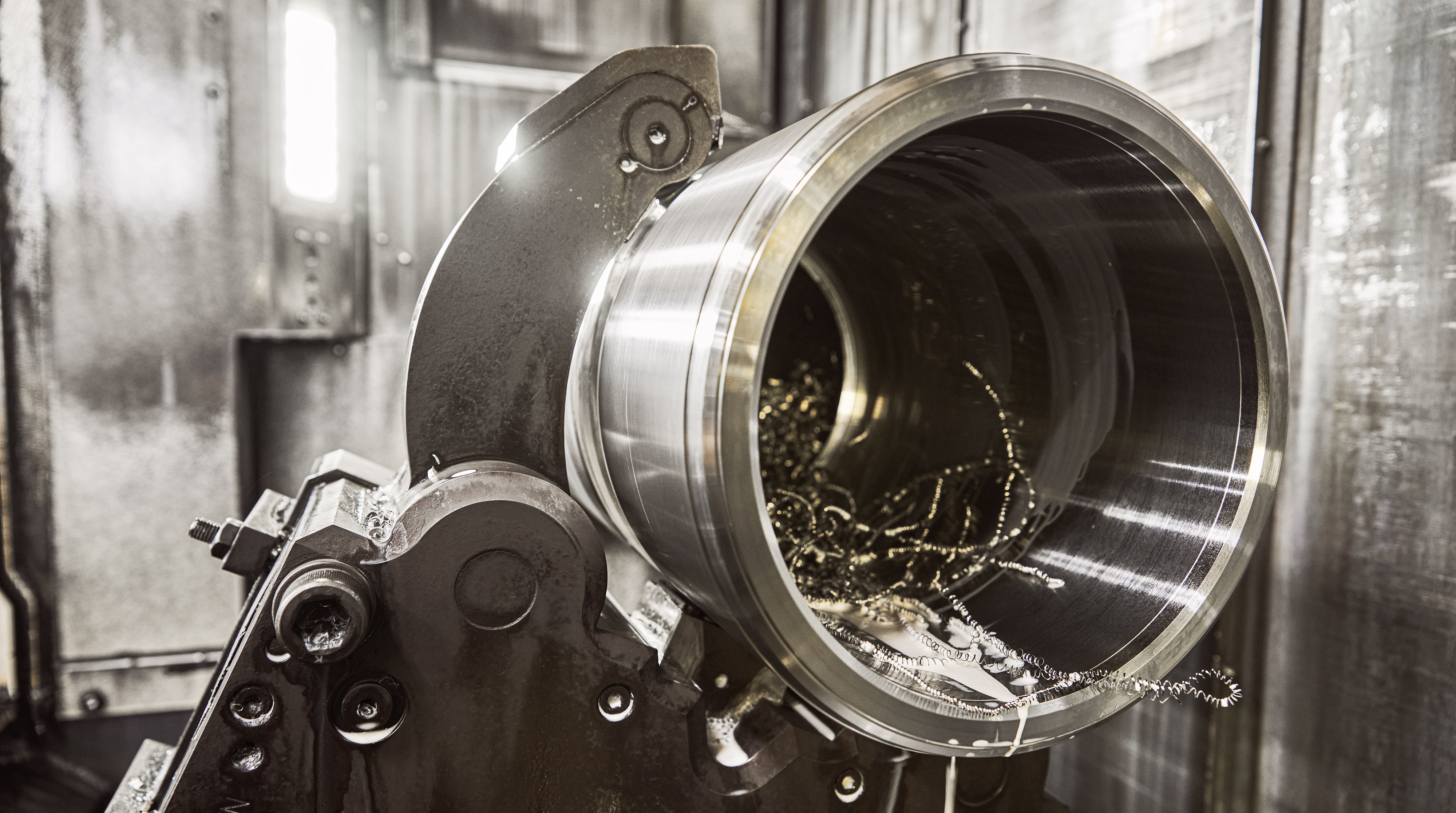 Nahaufnahme eines großen Metallzylinders, der bearbeitet wird, mit Metallspänen im Inneren. Die industrielle Umgebung ist schwach beleuchtet und metallisch.