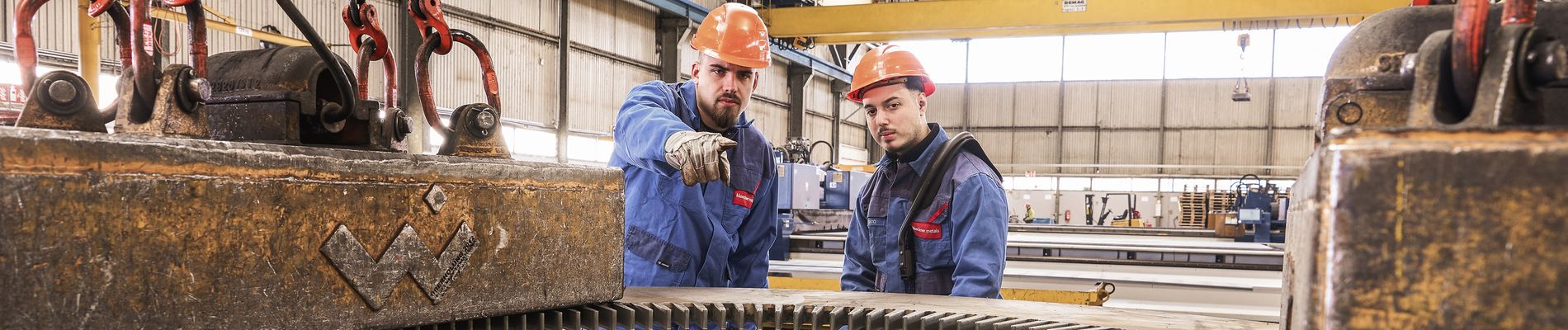 Zwei Arbeiter in orangefarbenen Schutzhelmen und blauen Overalls, von denen einer auf etwas zeigt, inspizieren ein großes Industriegetriebe und einen angehobenen Metallblock.
