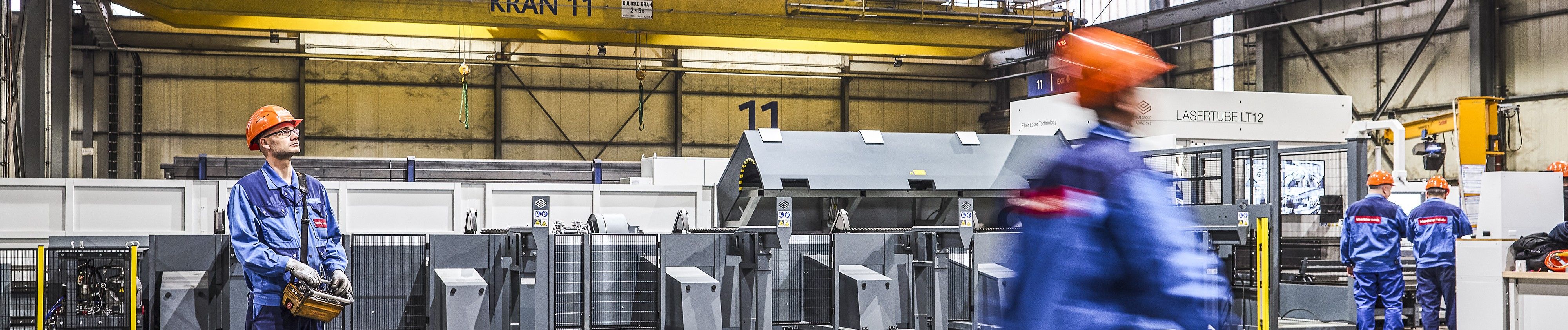 Industrielager mit Arbeitern in blauen Overalls und orangefarbenen Schutzhelmen um schwere Maschinen herum; einer hält eine Steuerbox in der Hand, während ein anderer verschwommen im Gehen zu sehen ist.