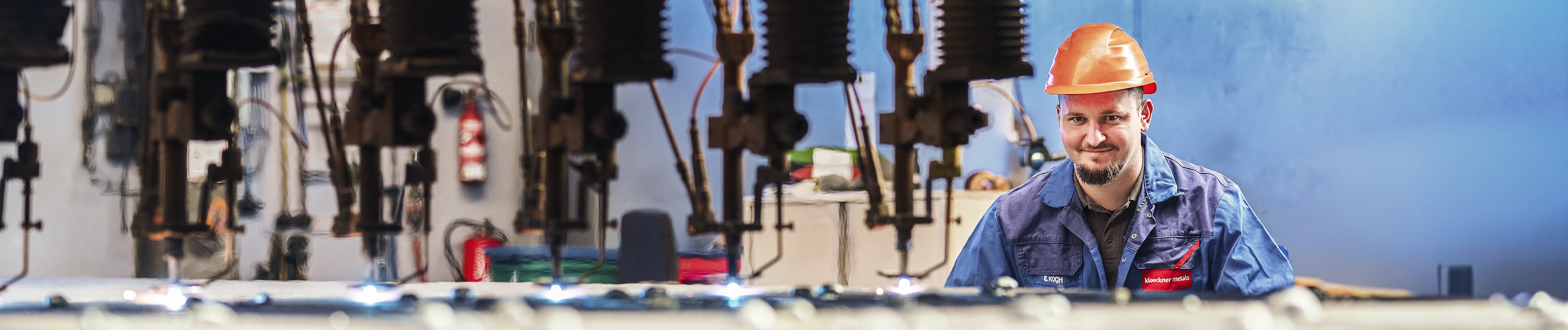 Ein Arbeiter in blauer Uniform und orangefarbenem Helm steht in einem Service Center, umgeben von Maschinen, und lächelt in die Kamera.