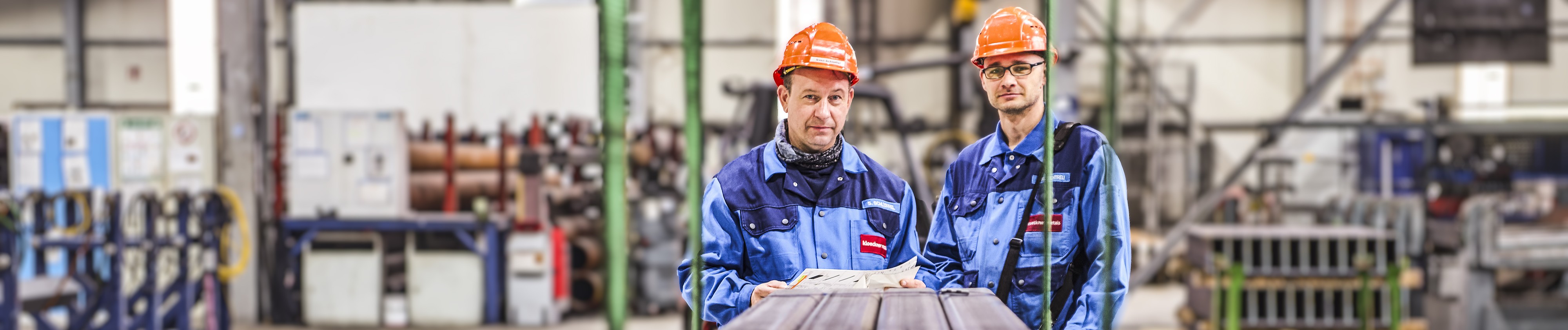 Zwei Arbeiter in blauen Overalls und orangefarbenen Schutzhelmen, die Papiere in den Händen halten, hinter ihnen verschwommene Maschinen.