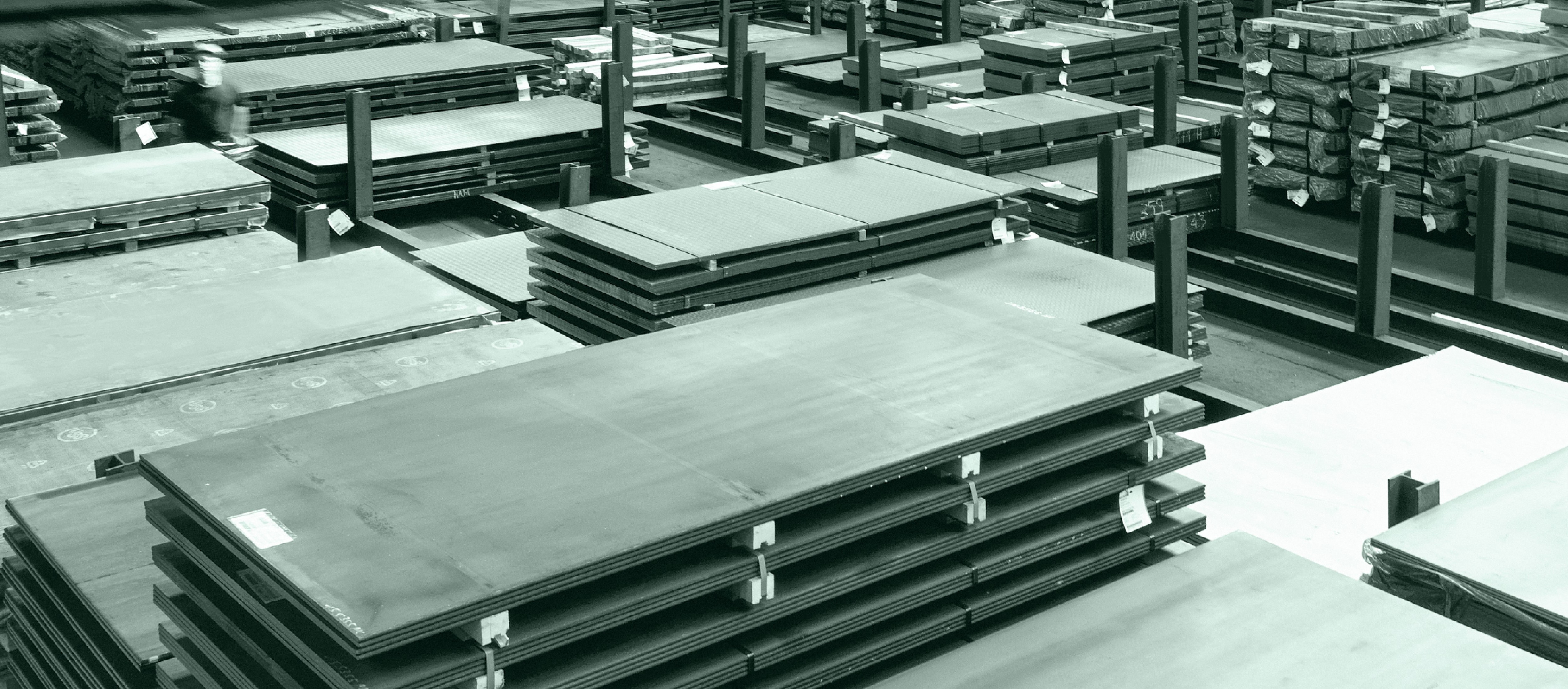 Stapel großer Metallbleche, die in einem Lagerhaus organisiert sind, mit Regalreihen und zusätzlichen Materialien im Hintergrund.