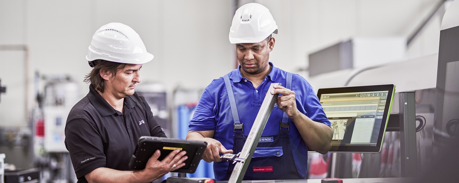 Two workers in hard hats operate machinery, one holding a tablet and the other adjusting a metal component, with a screen displaying data nearby.