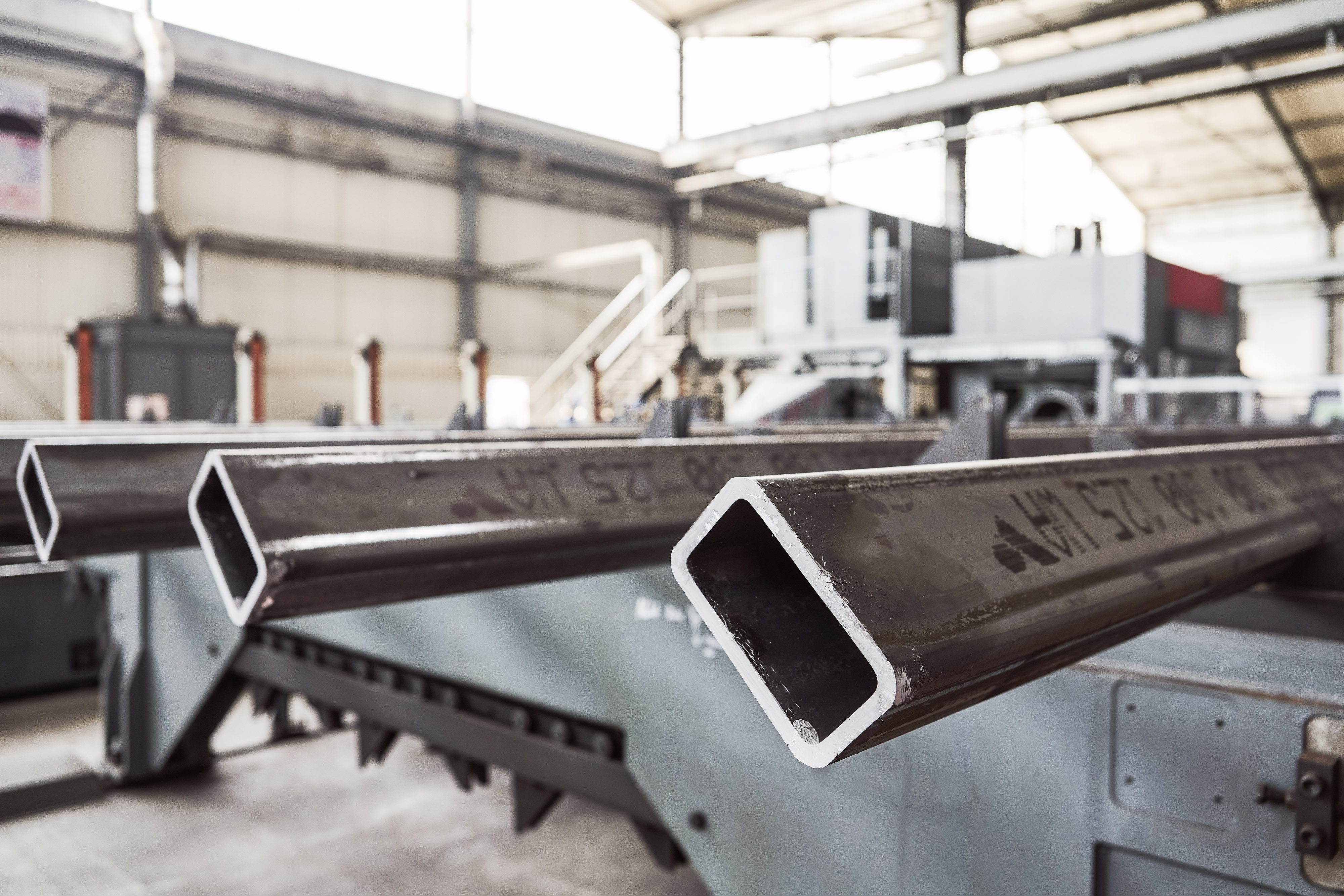 Close-up of metal square tubes in an industrial warehouse, with machinery and a high ceiling in the background.